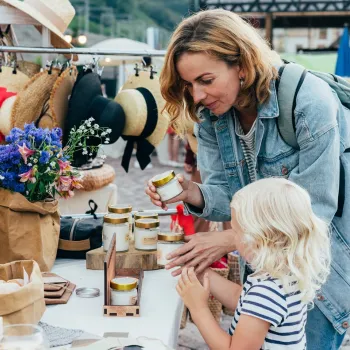 Moeder en kind bekijken handgemaakte producten op een zomerse braderie, met hoeden, bloemen en kaarsen op de kraam.