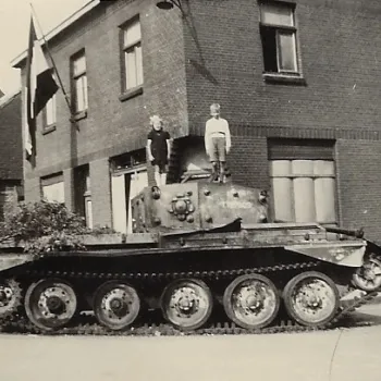 Zwart-witfoto van een beschadigde tank op een straathoek in een Nederlands dorp, vermoedelijk uit de Tweede Wereldoorlog. Twee kinderen staan bovenop de tank. Aan de omliggende huizen hangen vlaggen, wat wijst op een bevrijdingsmoment.