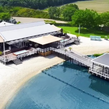 Luchtfoto van Beachclub Degreez in Panheel, waar het weddingevent plaatsvindt, gelegen aan het water met een zandstrand, houten terrassen en een steiger die uitkijkt over het meer, omringd door groen landschap.