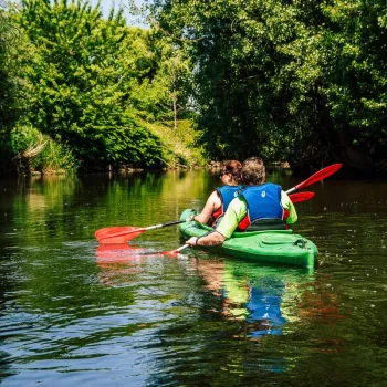 Two people canoeing a green kayak along a peaceful, tree-lined river.